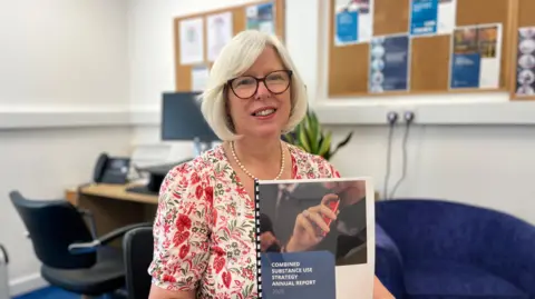 BBC Dr Nicola Brink holding the Public Health's Combined Substance Use Strategy 2025 Annual Report in her office. She has medium length white hair and glasses, with a floral dress on.