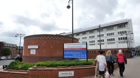 PA Media Main entrance to Leeds General Infirmary - with a circular brick building with a large sign reading Leeds Teaching Hospitals NHS Trust and directions to parts of the site. There is a large white building behind.