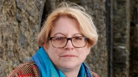 Prof Marion Gibson, sitting outside a historic stone building. She has fair, centre-parted hair and is wearing tortoiseshell glasses and a turquoise scarf over a thick red, black and brown checked coat.