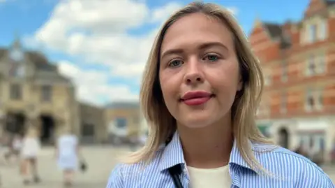 Daisy Blakemore-Creedon vestindo uma camisa listrada azul e branca sobre uma camiseta branca sorri para a câmera enquanto é fotografada do lado de fora de um prédio.