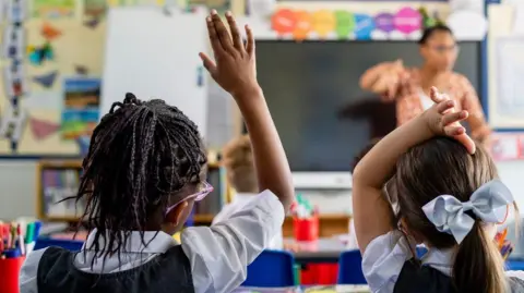 Two children with their hands raised in a classroom setting. The image is from the back of their heads. One has black braids in and is wearing pink/purple framed glasses and the other has brown hair tied in a ponytail with a blue bow. Both are wearing light short sleeved shirts and dark pinafores. 