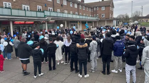 Kate Bradbrook/BBC A large group of people holding blue balloons, the crowd seen from behind. They are outside a shopping precinct. Adults and children are together.