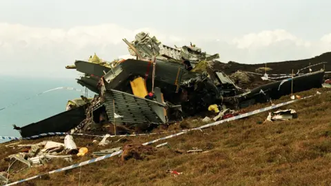PA Media A photograph of the wreck of the Chinook helicopter, which had crashed in Scotland. It is on a large mound of grass, with police tape placed around the crash site.