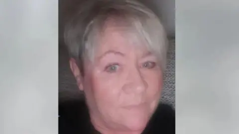 Kelly smiles a small smile in a selfie. She is sat on a sofa and has short grey hair. 