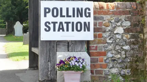 A sign which says "Polling Station" is attached to a wooden bench and old brick wall. There is also a lilac flower pot with purple flowers. 