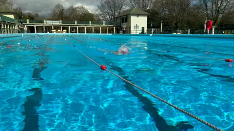 Emma Baugh/BBC Peterborough Lido, which has separate swimming lanes. A single swimmer is in the pool. The sky is cloudy. The water is bright blue and there is a lifeguard dressed in red at the side of the outdoor pool.