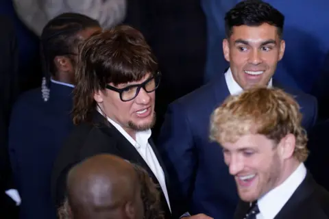 Getty Images A antheral   with a mullet and glasses stands adjacent  to a antheral   with acheronian  hairsbreadth  and a antheral   with blond shaggy hair. They each  deterioration  suits.