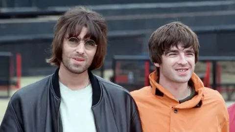 Liam and Noel Gallagher smiling together in front of the stage at Knebworth House during the day