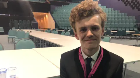 Emma Baugh/BBC Sam Carling, in a jacket and tie, smiling in an empty hall where the election count was held.