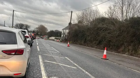 A long queue of traffic lines a road marked with orange traffic cones.
A white car sits in front of a red car as vehicles wait in Ballykelly.