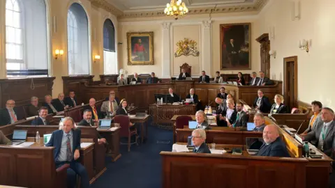 BBC There are 38 politicians sitting at wooden benches all looking beyond the camera in Guernsey's Royal Court