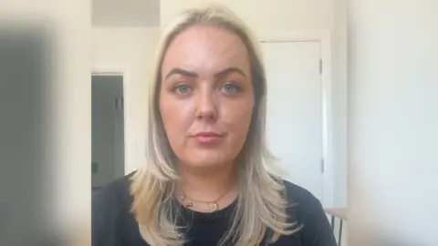 A woman with shoulder length blonde hair, wearing a black tshirt, staring at the camera expressionless