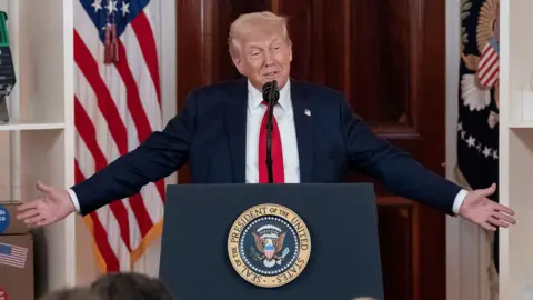 Donald Trump pictured speaking wearing a blue suit and red tie at an event at a podium marked with a circular White House logo. Behind him, there are objects as part of an event highlighting business investment in the US 