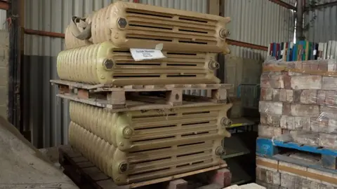 Martin Giles/BBC A pile of dull gold-coloured radiators are stored on their side on pallets, next to pallets of bricks in a corrugated iron building.