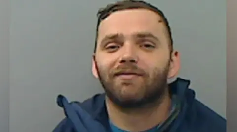 A man with short brown hair, a brown beard, smirks at the camera, in a police mugshot. The background is a painted grey wall and he is wearing a blue t-shirt and navy jacket. 