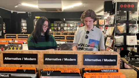 Hermione and Max browse records in a shop. The names of music genres plus acts like Supergrass and Jamie XX can be seen on cards above the records.
