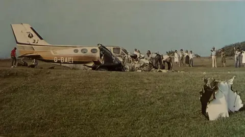 An old photograph shows the wreckage of a small aircraft in a grassy field while people dig through its remains and others watch on.