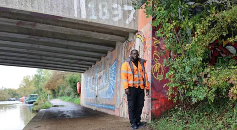 Devon standing by a canal on an overcast day. He is wearing an orange vis jacket. There is greenery growing near him. There is graffiti on the wall behind him. A boat can be seen moored in teh distance.