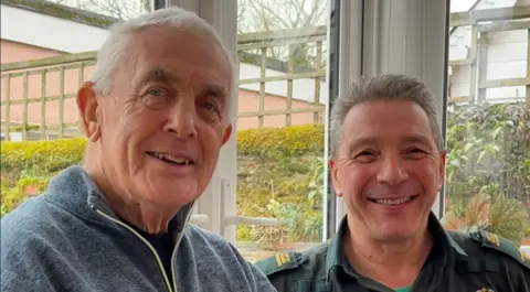 A head and shoulders image of Ted Tracey, wearing a zip-up grey top, next to Gary O'Shea, in a green shirt that is his first responder uniform. Yellow lettering on the epaulettes is too small to be read. Both men have grey hair and are smiling at the camera, and in the background there are windows and French doors and a view out to a garden fence.