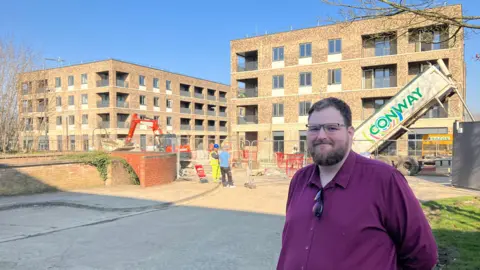 Amy Holmes/BBC A man, councillor Ed Hume, stands in the foreground near a construction site with new brick apartment buildings, while workers and machinery operate behind safety barriers.