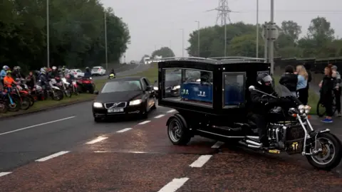 A trike motorcycle carrying Layton Carr's coffin turns left off the main road. The coffin, coloured blue, can be seen through glass windows. A number of bikers are parked on a nearby grass verge.