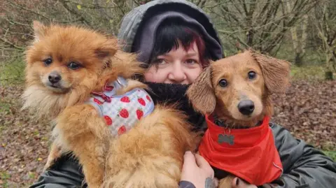 Lizzy's Pet Services A woman, with black and red hair, with a coat on and its hood up, holding onto two dogs, in each arm. She is looking at the camera and so are the dogs. Trees are behind her and leaves are on the ground. Both dogs have coats on. 