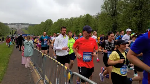 Thousands of participants run along Prince of Wales Avenue, the mile-long tree-lined drive through the Stormont estate. There are metal barriers along the route, separating runners from spectators. There is a dull, grey sky and the grey facade of Parliament Buildings can be seen in the background. 