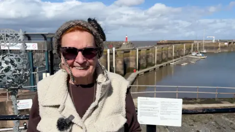 Loretta Whetlor wears sunglasses and a white fluffy gilet over a brown top. She is standing in front of a harbour area on a sunny day with some clouds.