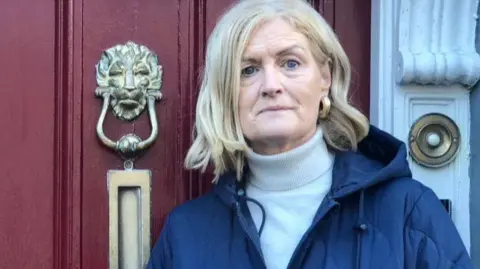 Kathleen Feni stands outside the front door of her house. The door is red with a gold letterbox and gold knock. There is a white column on the right side with the gold door. Kathleen has golden hair and is wearing a cream polonac and blue coat. She is wearing Gold Hoop earrings.