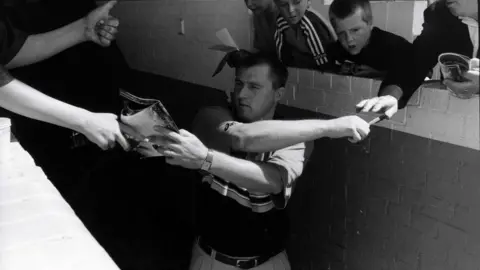 Ian Beesley A black-and-white photo of a man in a polo shirt crossing his arms as he tries to sign a football programme while giving back a pen to a young football fan.