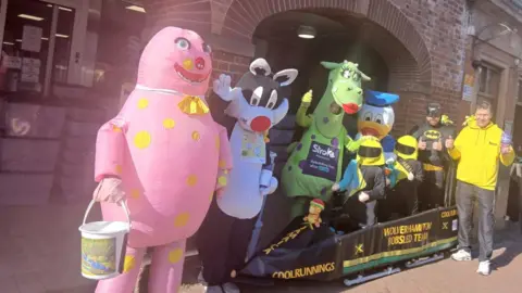 The group fundraising in fancy dress. From L-R, the group is dressed as Mr Blobby, Sylvester, Puff the Magic Dragon, Donald Duck and Batman. In front of them, two of the group are in the bobsled wearing helmets. 