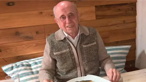 Norman Irwin is bald with some grey hair on the side of him. He is wearing a brown patterned zip-up fleece with a white shirt beneath it. He is wearing metal glasses and is smiling at the camera as he sits down with a wooden wall and striped cushion behind him.