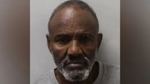 Police custody photograph of Musse Ali, a man with short grey hair, a short grey beard, and a grey crew-neck top.