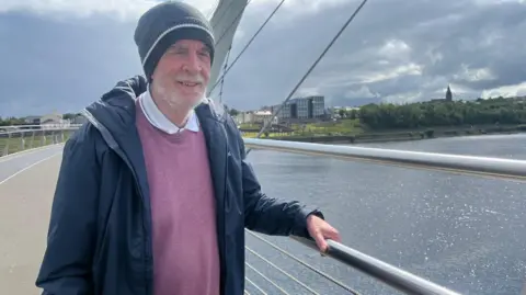 a man stands on a bridge wearing a blue jacket and pink jumper. he has a grey hat on his head. a river is behind him, and a number of buildings can be seen in the background