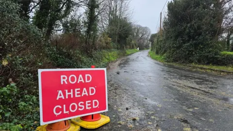 A red road closed sign on the side of a very wet country lane.