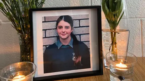 McEgan College A framed photograph of Lauren who is wearing a navy school jumper and has a grey shirt collar. She has very long brown hair in a ponytail that is coming over her shoulder.