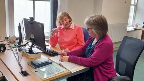CAB Two women in front of a computer screen looking at book on a desk. One is wearing a coral jumper and holding her glasses. The other has a blue dress and pink cardigan and is holding a pen. 
