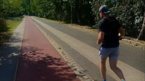 Angus Read A man in blue shorts, a black tshirt and grey cap running along an empty sunny road