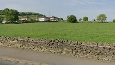 A large green field contained by a dry-stone wall. In the background, residential houses can be seen.