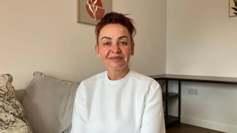 JOANNA TAYLOR/BBC Kelly Slack is smiling and looking at the camera. She is wearing a plain white t-shirt and multiple earrings. Behind her are large grey cushions and a white wall with a framed print of a plant on it.