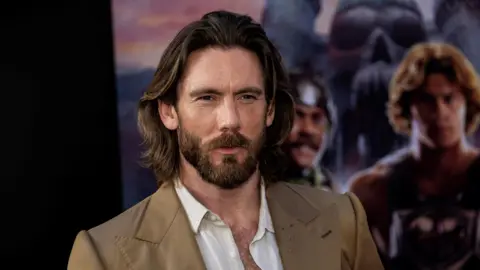 James Wilkinson. He has long dark hair and a beard. He is wearing a tan suit jacket and an open white shirt. A blurred movie poster is behind him. 