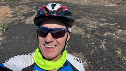 A selfie of a man wearing a red, white and black cycle helmet and sunglasses, along with a blue, black and white lycra top. Behind him is a patch of what looks like wasteland, with a few small buildings in the background.