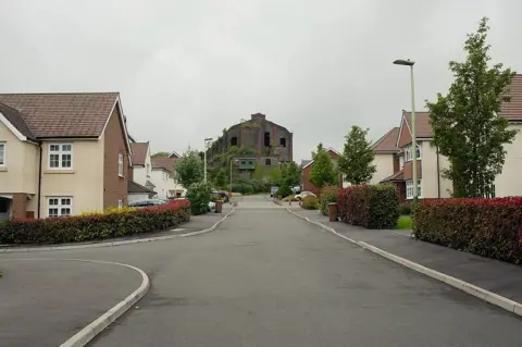 Jon Pountney Houses with a stone factory building with plants growing on it at the end of the street