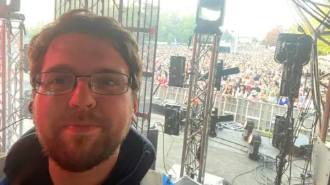 RICHARD WALPOLE An early thirties male with a beard and glasses, standing back stage at a festival in front of a large crowd.