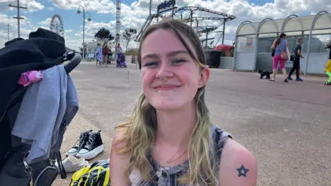 A woman with long blonde hair smiles at the camera. She is sitting next to a pram, a bag and some pairs of shoes. She is wearing a black and white checked, sleeveless top and two tattoos on her left arm. Behind her are fairground rides.