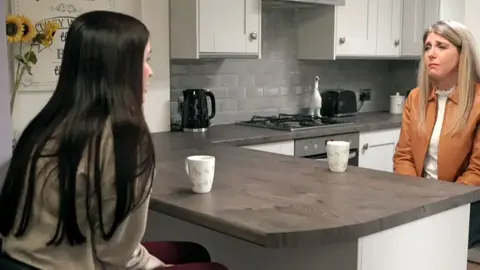 Leanne Lucas sits across from reporter Judith Moritz. They are in a kitchen and both have white mugs in front of them. Leanne's long black hair is down her back and she wears a grey top. Judith wears a dark orange jacket.