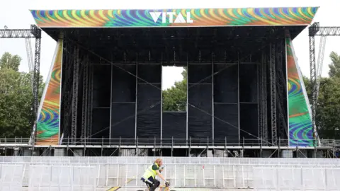 Image of the Vital stage set up at Boucher Road Playing Fields, a man working is riding his bike passed the stage. He has a hi vis jacket on. The stage is metal and has a sign reading VITAL on the top of the stage. 