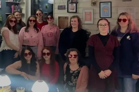 Analise Taylor A group of young women, some of whom wear red heart shape sunglasses, smile and pose for a photo in a bar.