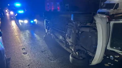 A white car on its side after a crash in a residential road. There are both a police car and an ambulance behind it with blue lights on. It is dark.