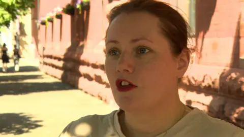 Elaine has brown hair tied back and is wearing a cream coloured top. Her mouth is slightly open, as though talking.